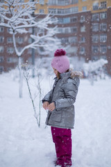 Little girl in a silver jacket and a hat with a pompom walks in the park near the house among the snow covered winter trees