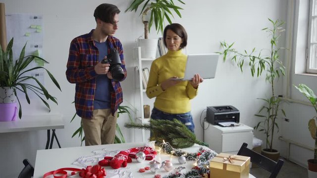 Male photographer taking Christmas flatlay images with camera while female stylist or assistant checking pictures on laptop and arranging objects on table