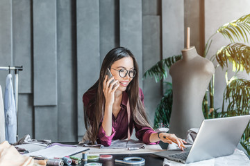 Young asian fashion designer using laptop and calling by phone in her studio