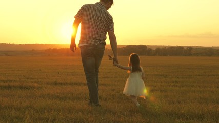 little daughter walks with her father in meadow holding hands. child holds father's hand. family walks in evening out of town. dad and baby are resting in the park. child plays with his father