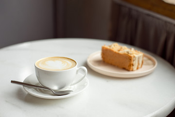 Slice of caramel cake and a fork on the right side. A cup of hot coffee with ground in the background.Close-up