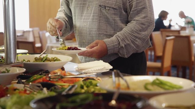 American man puts tasty food on plate while standing in restaurant buffet. Male client serves himself, choosing varied cooked meal for dinner. People eating and sitting at table in interior. Concept