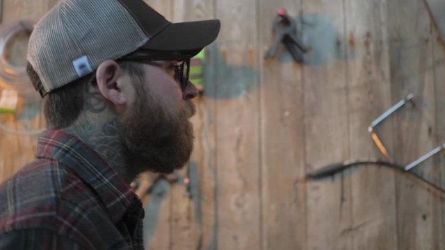 Male walking across old fashion rustic warm tool shed work space with equipment hanging on wooden walls with beard hat glasses and plaid shirt in slow motion
