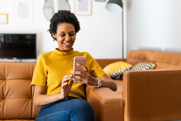 Smiling young woman sitting on couch at home using smartphone