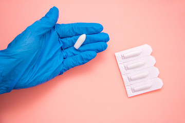 White suppository for anal or vaginal use in hand of a doctor wearing gloves on a pink background....
