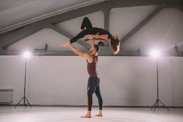 The theme of Acroyoga and Yoga Poses. A pair of two men and a woman stand in the position of asana. The guy holds the girl arched high back on the outstretched arm. In the gym with a studio Backlight