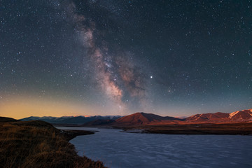 Beautiful starry night landscape. The bright galaxy of the Milky Way over a frozen lake and orange mountains lit by the light of sunrise.