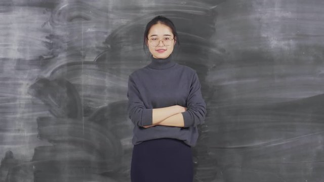 Lockdown of attractive young Asian woman standing against cleaned blackboard, turning around, crossing arms on her chest and looking at camera