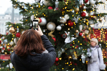 Woman photographing her girlfriend by smartphone on Christmas tree background. New Year decorations and festive lights on a city street