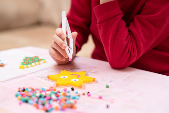 Little girl holds a tweezers with bead, creates art toy from fusible colorful beads, last touches. Also known as perler beads. Toy that develops the imagination of child. Process close up
