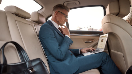 Business trip. Side view of handsome mature businessman in full suit working on his laptop while sitting in the car