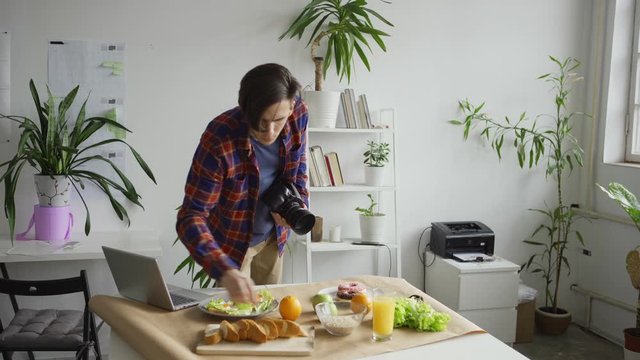 Medium shot of young male photographer arranging objects while taking food background pictures with fruits and meals placed on table in office