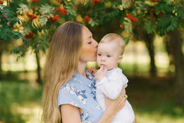 A sweet and gentle young woman a European mother holds her little son in her arms and kisses him near the branches with red Rowan