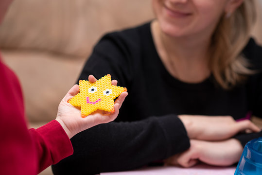Little girl shows mom just crafted art toy from perler beads, beautiful smiled star. Fusible colorful beads. Toy that develops the imagination and creativity of child