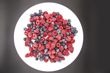 A Berry mix  from frozen raspberries and blueberries on the white plate. A Frozen Berries in black background.  A sweet background with frozen raspberries and blueberries