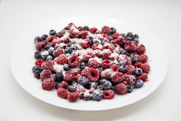 A Berry mix in sugar from frozen raspberries and blueberries on the white plate. A Frozen Berries with Sugar.  A sweet background with frozen raspberries and blueberries