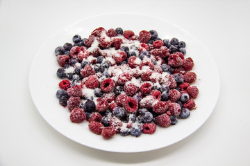 A Berry mix in sugar from frozen raspberries and blueberries on the white plate. A Frozen Berries with Sugar.  A sweet background with frozen raspberries and blueberries