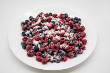 A Berry mix in sugar from frozen raspberries and blueberries on the white plate. A Frozen Berries with Sugar.  A sweet background with frozen raspberries and blueberries