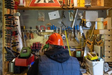 Craftsman with a construction helmet on the phone with customers, sitting in front of a workbench overfilled with rubbish