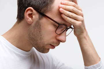 Fototapeta premium Close up portrait of man in glasses in white top touching his forehead, feels tired after working on laptop, white background. Overwork, exhausted, chronic fatigue, mental stress, headache concept. 