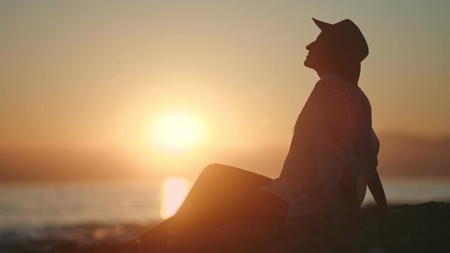 Silhouette of relaxed tourist woman in hat contemplating amazing natural sunset sitting on coast