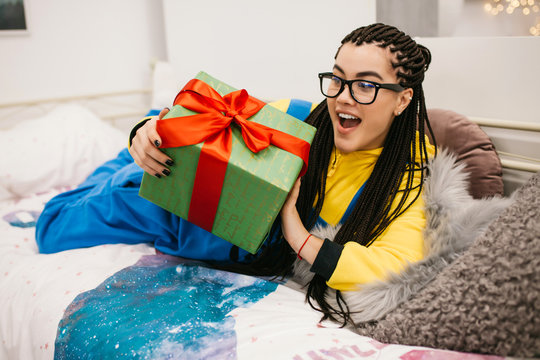 Happy birthday or happy New Year! Cheerful girl with afro braids in minion pajama holding birthday gift box sitting on a bed and smiling. Crazy emotions. Pajamas party
