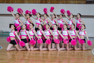 attractive young cheerleaders working out in sports club, group of cheerleaders with pom-poms in their hands, hands raised up, girls in black and pink suit with pompons on the background of the gym