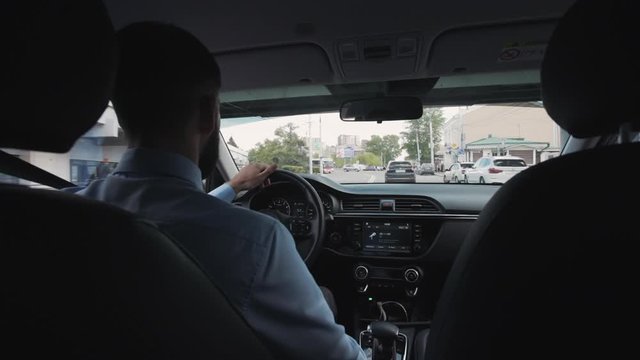 Back View Of Caucasian Adult Businessman Driving Car In Summer Day. American Man Driver Holding Steering Wheel In Hand, Watching Away In Windshield Sitting In Black Interior Of Auto. Handsome Guy