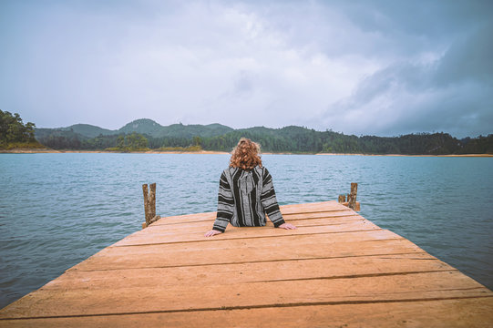 Young woman sitting on a pier watching a lagoon with mountains in the background and cloudy sky