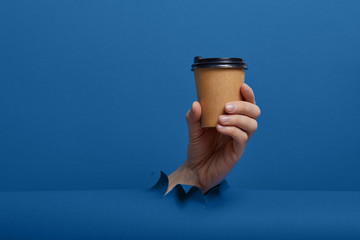 Male hand taking a cup of coffee through torn classic blue paper background. Get energy boost, hot drink in take-away package.