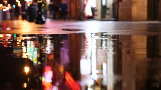 People walk through puddle in night city, lighten by neon signs.