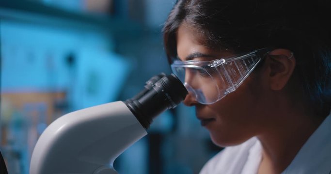 Female research scientist looking into microscope, wearing safety glasses.Blue lighting in a dark lab room.Close up, slider, shot with BMPCC 4K.Biochemistry, pharmaceutical medicine, science concept