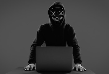 Anonymous man in a black hoodie and neon mask hacking into a computer