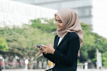 happy expression of beautiful young asian muslimah business woman on black suit sitting outside using mobile phone