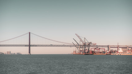 An evening on the Tagus river in Lisbon, Portugal: suspension bridge "Ponte 25 de Abril", recognized as the most beautiful bridge in Europe, commercial docks with cranes and cargo containers