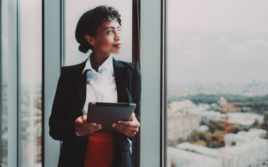 A dazzling young African-American woman entrepreneur with a digital tablet in her hands is standing near a huge panoramic window indoors of a business high-rise and pensively looking at the cityscape - Powered by Adobe