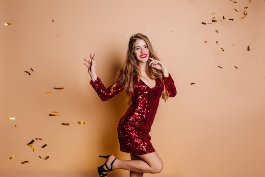 Lovely slim girl in red sparkle party dress dancing with confetti and laughing. Indoor photo of elegant young woman in black shoes and hair accessory relaxing during new year event.