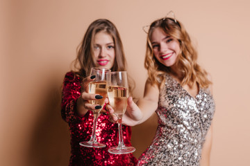 Indoor blur portrait of spectacular young ladies with wineglasses in focus. Pretty girl with blonde curly hairstyle celebrating birthday with her best friend in red dress.