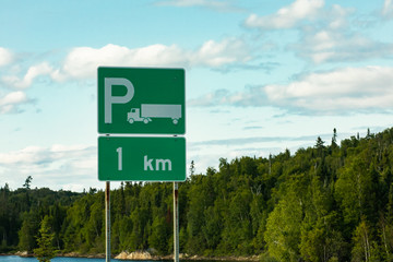 Trucks Parking Green Sign with a white long truck symbol, after 1 km, with lake, pine trees, and cloudy sky in the background