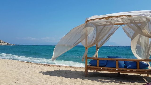 Beautiful light outdoors Sheer white curtains of the modern wooden gazebo fluttering on the summer breeze, white sand beach in Greece. Background shot with copy space