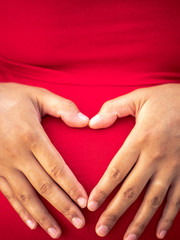 A young African American mixed race or ethnicity woman wearing a bright red dress holds her hands on her pregnant belly forming her fingers in the shape of a heart making a beautiful maternity photo.