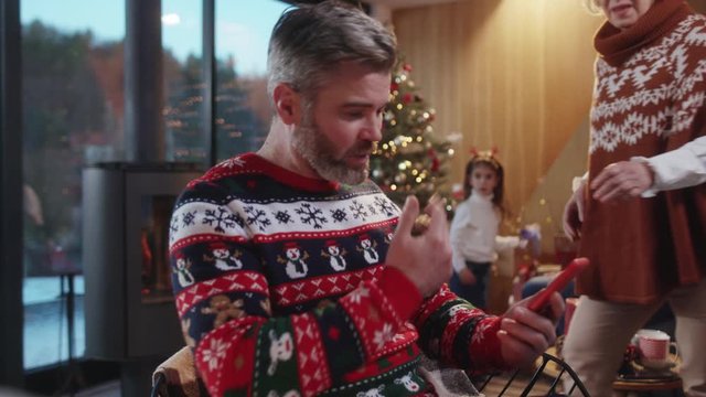Ecstatic bearded father in sweater reading news on smartphone calling his big family showing amazing holiday greeting hugging together in cozy living room. Christmas time.