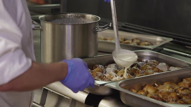 closeup. chef hand pouring baked chicken with white sour cream sause in buffet cafeteria steel container. cook preparing meat for catering public cafe. appetizing, canteen business concept