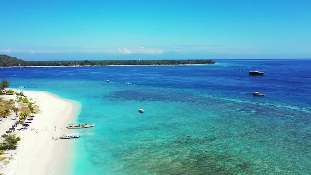 Paradise tropical bay with white sandy beach and blue turquoise calm lagoon where boats floating on a bright sky over low-lying island in Indonesia