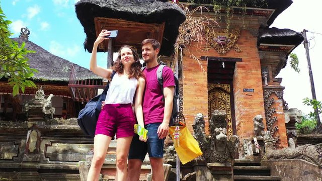 Happy young couple taking selfie photos with Asian ancient temple background full of decorated walls and stone statues 