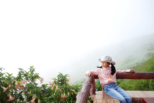 Young Asian girl in little cowboy costume (North plaid shirt, blue jeans and Sombrero hat) sitting on wood balcony with mountain landscape and sea of mist. Phutabberk, Petchabun, Thailand. Copy space.