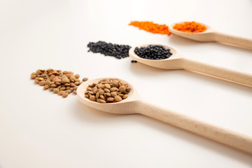 a wooden spoon with brown lentils close-up, in the background are two spoons with black and orange lentils out of focus. White background.
