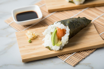 Lachs Fisch Avocado Temaki Sushi, eingelegter Ingwer mit Soja Soße und Sushimatte auf Bambus Brett Teller und Marmor Hintergrund