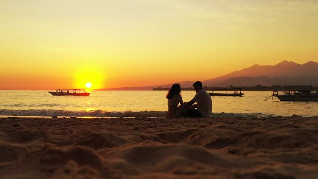 Romantic couple male and young woman silhouette sitting on the tropical beach enjoying sunset and ocean. Wind is playing with female long blond hair Bali Indonesia South East Asia