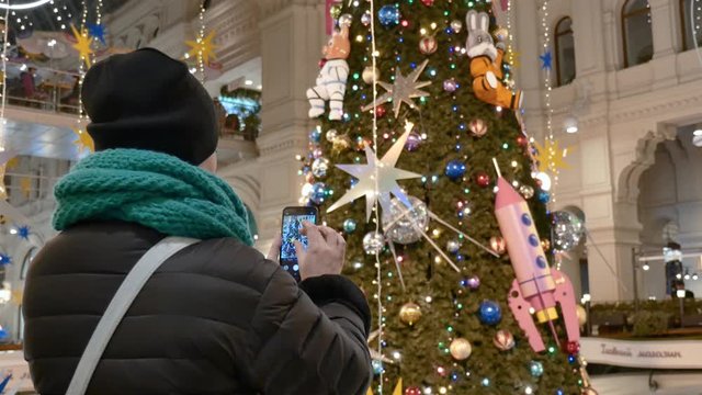 Caucasian woman takes photo of Christmas tree in shopping Mall. She is dressed in a black coat and hat, a green warm scarf. The view from the back. Close up